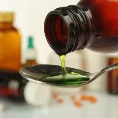 Zielony syrop prawoślazowy dla psa kapie z butelki na łyżkę. W tle widać inne leki.